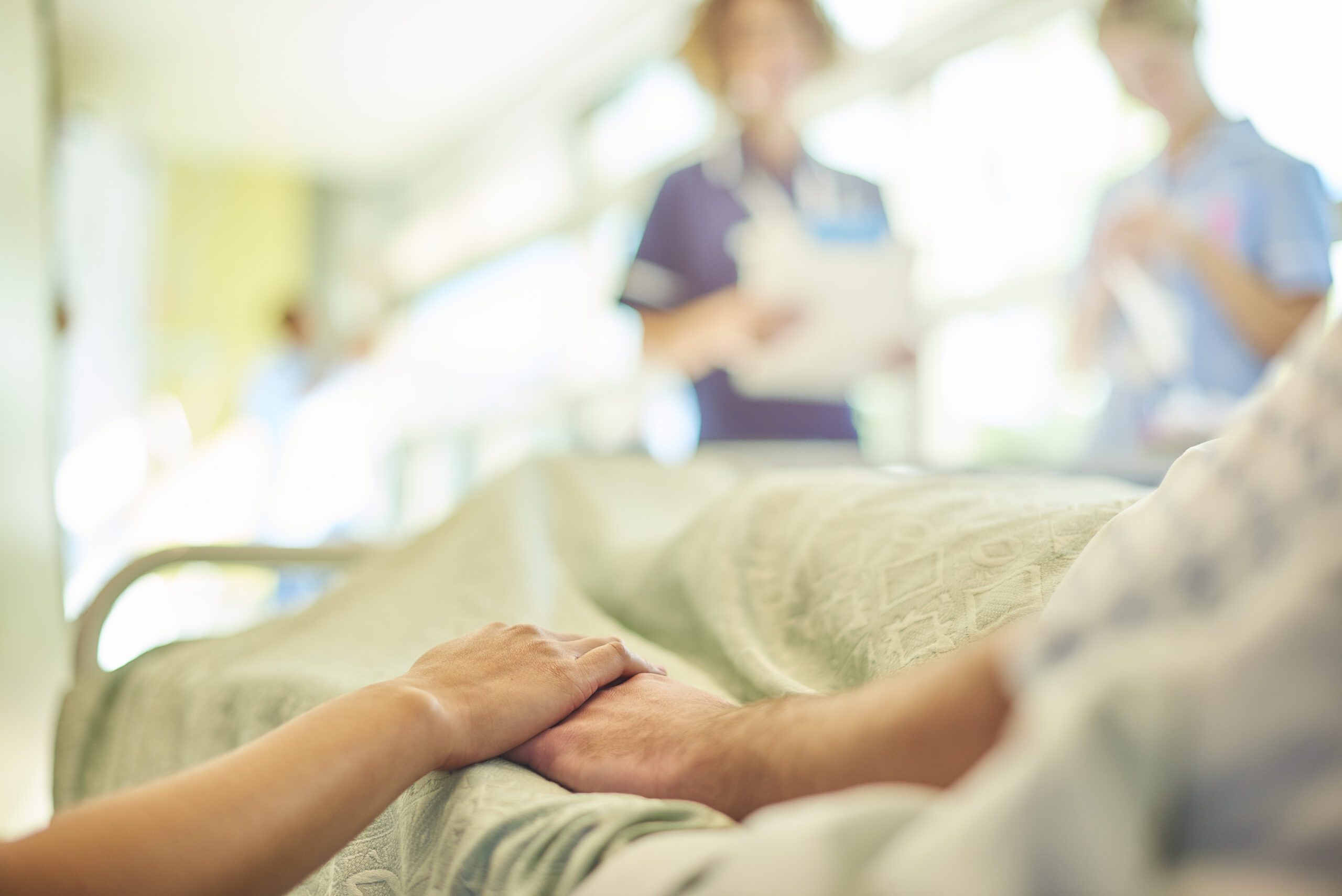 a hospital visitor's hand holds a patient's hand in bed of a hospital ward. In the blurred background a young nurse is chatting to the ward sister about the patient's care.